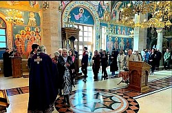 Верници и свештенство окупљени у централном делу Храма Преображења Господњег на Златибору током дугог братског састанка. Један свештеник врши чин благосиљања или исповести верника, док се у позадини види карактеристична мермерна розета на поду и бочни олтар са дрвеним елементима и иконама