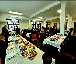 Сабор свештенства намесништава ужичког и рачанског за заједничком трпезом љубави у пространој сали парохијског дома Храма Преображења Господњег на Златибору након братског састанка. Дуги столови су постављени са посном храном, рибом, салатама и воћем, док свештеници у црним мантијама разговарају и заједничаре у светлом амбијенту