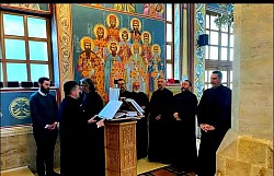 Свештеници Жичке епархије у првом плану поју за певницом током Литургије пређеосвећених дарова у Храму Преображења Господњег на Златибору. Кадар наглашава молитвени израз и заједништво свештенства током братске исповести и састанка намесништава ужичког и рачанског. У позадини се назире унутрашњост златиборског храма са богатим дуборезом и фрескописом